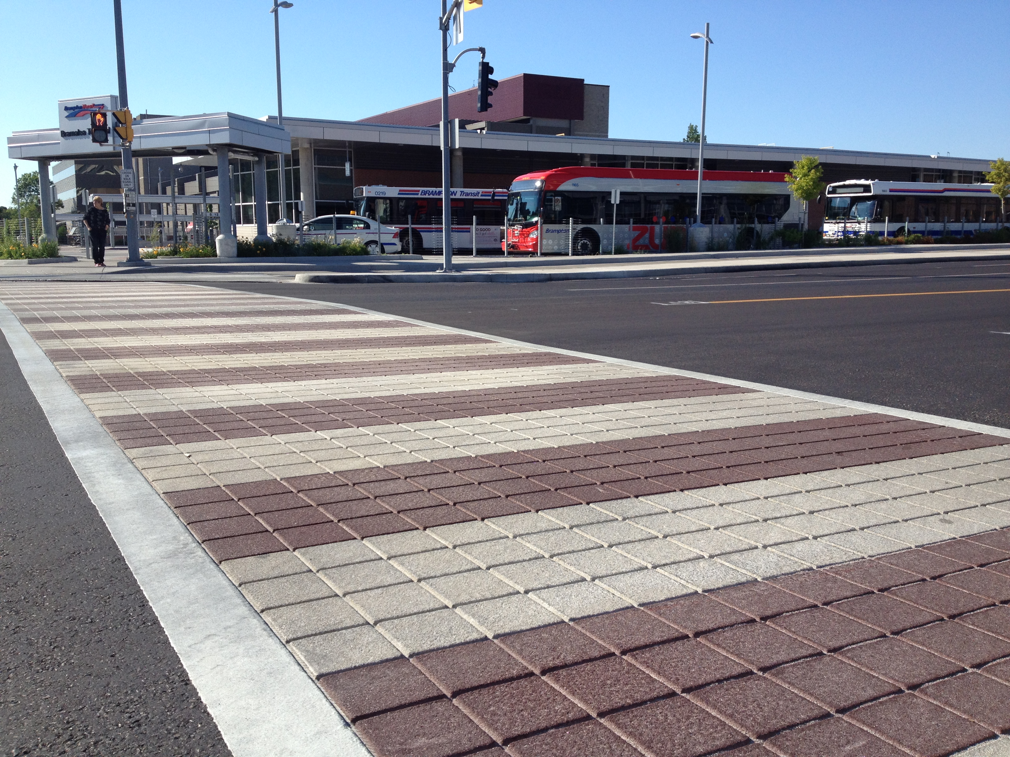 TrafficPatternsXD crosswalk in alternating red and pale brick-pattern bands at a bus terminal, buses at the platforms behind