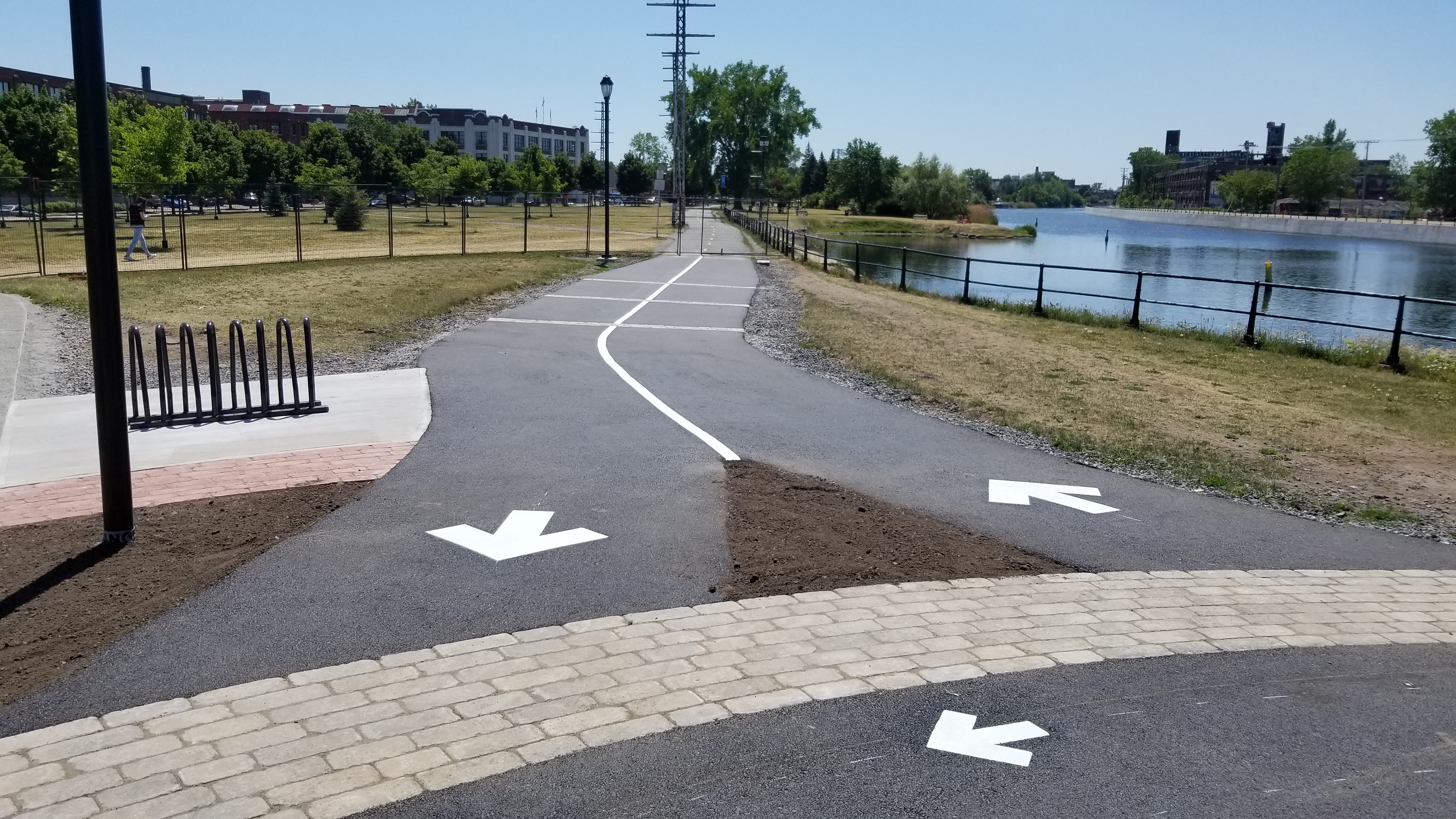 Waterside multi-use path with white direction arrows and a curving centre line, bike racks and a paver edge in the foreground