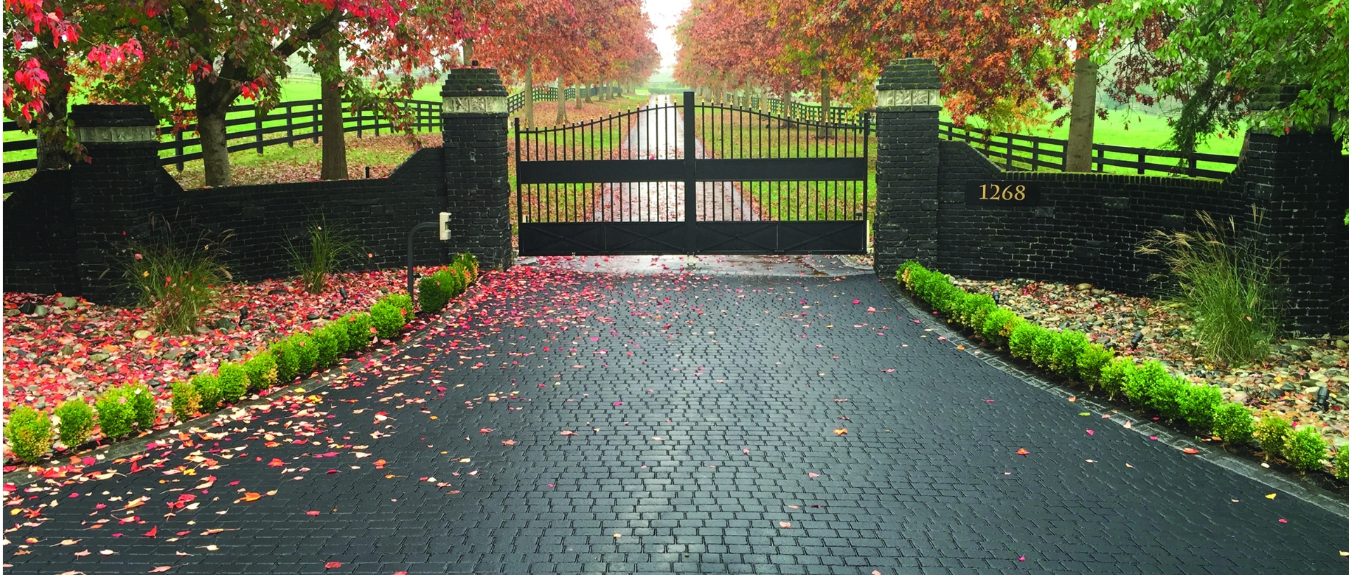 StreetPrint stamped asphalt estate driveway: dark cobblestone-pattern entry between brick gate pillars under autumn trees